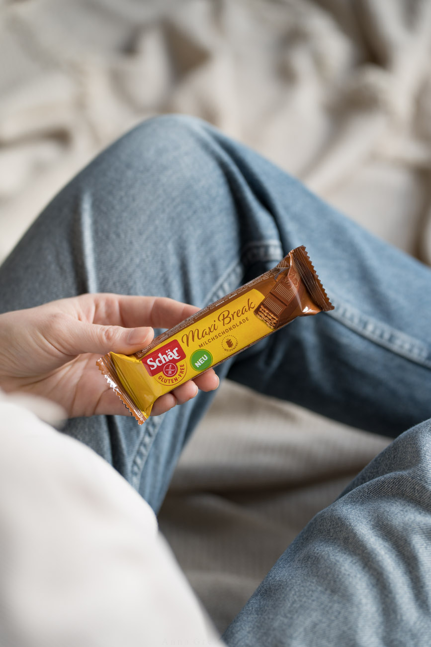 Eine person sitzt auf einer beigefarbenen Decke und hält einen Schokoriegel in einer gelben Verpackung in ihrer Hand. Das Foto wurde über die Schulter aufgenommen, dass man nur die Beine und den Riegel sieht.