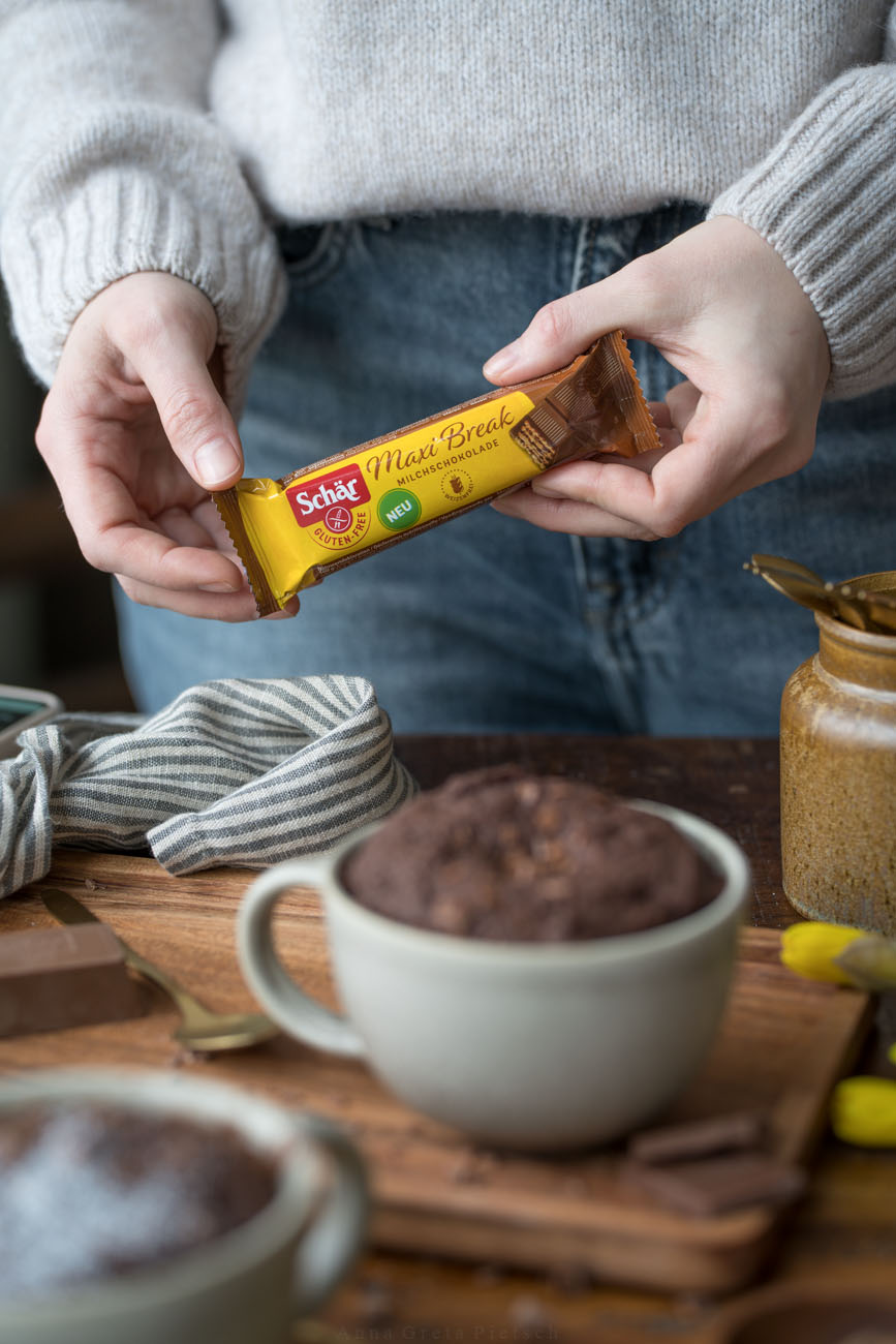 Eine Person steht hinter einem Holztisch und hält einen Schokoriegel in einer gelben Verpackung in den Händen. Vor ihr steht eine große Tasse mit einem Schoko-Tassenkuchen.