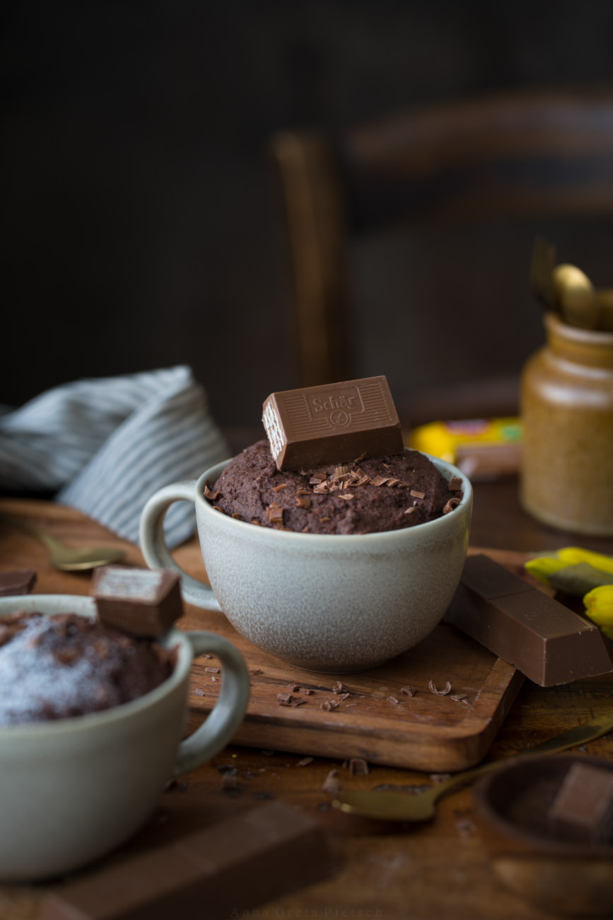 Man sieht einen Holztisch, darauf steht in der Mitte eine große Tasse mit einem Schoko-Tassenkuchen. Darauf liegt ein knuspriger Riegel.