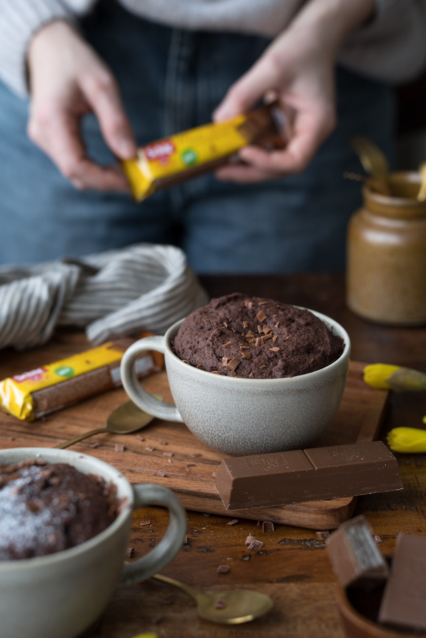Ein Holztisch, darauf steht in der Mitte eine große Tasse mit einem Schoko-Tassenkuchen. Eine PErson steht dahinter und hlt einen Schokoriegel in einer gelben Verpackung in den Händen.