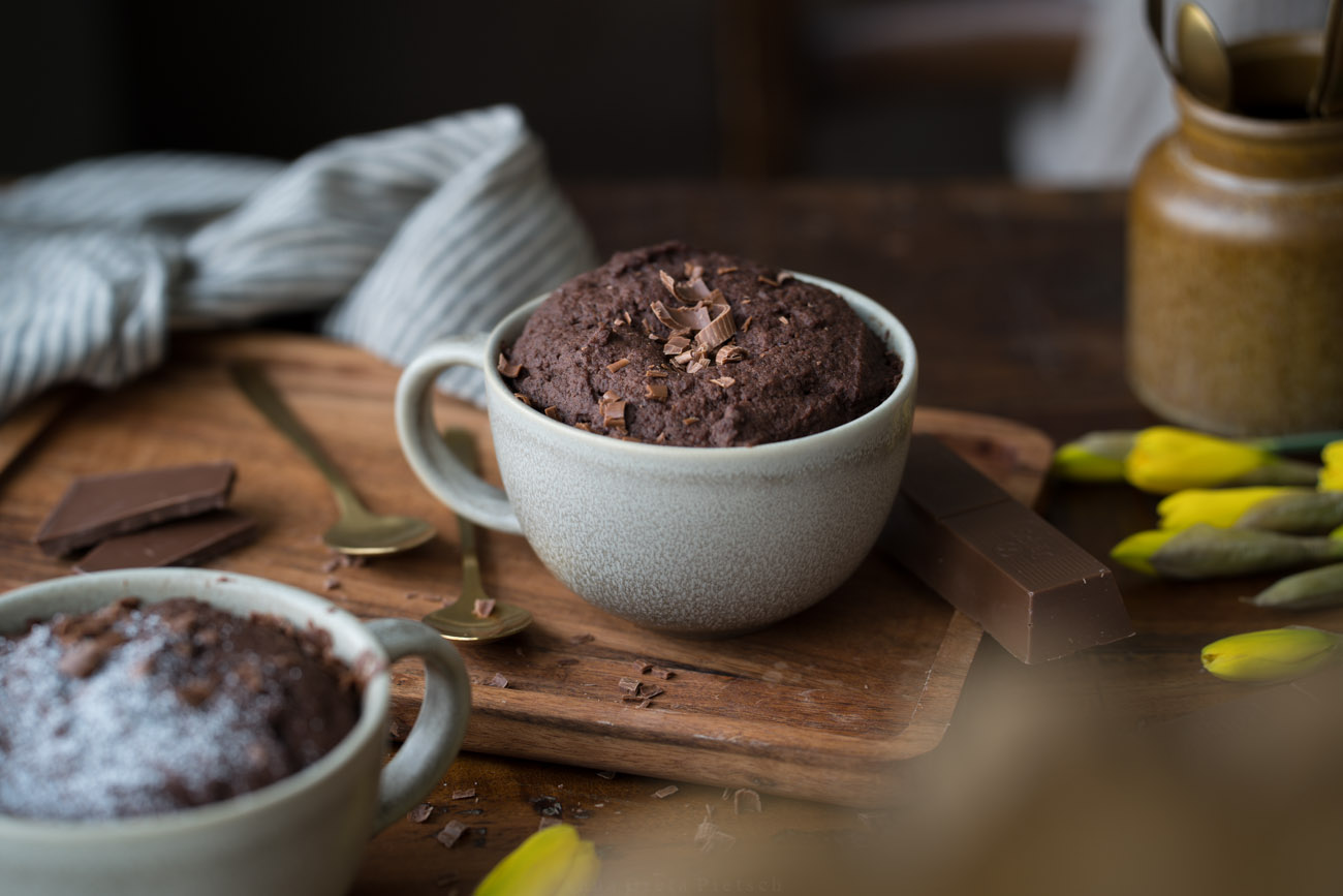Man sieht einen Holztisch, darauf steht in der Mitte eine große Tasse mit einem Schoko-Tassenkuchen.