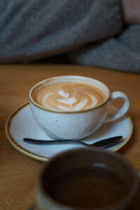 Cappucchino in einer großen, hellen Tasse. Sie steht mit einem Unterteller auf einem holzfarbenen Tisch in einem Café.