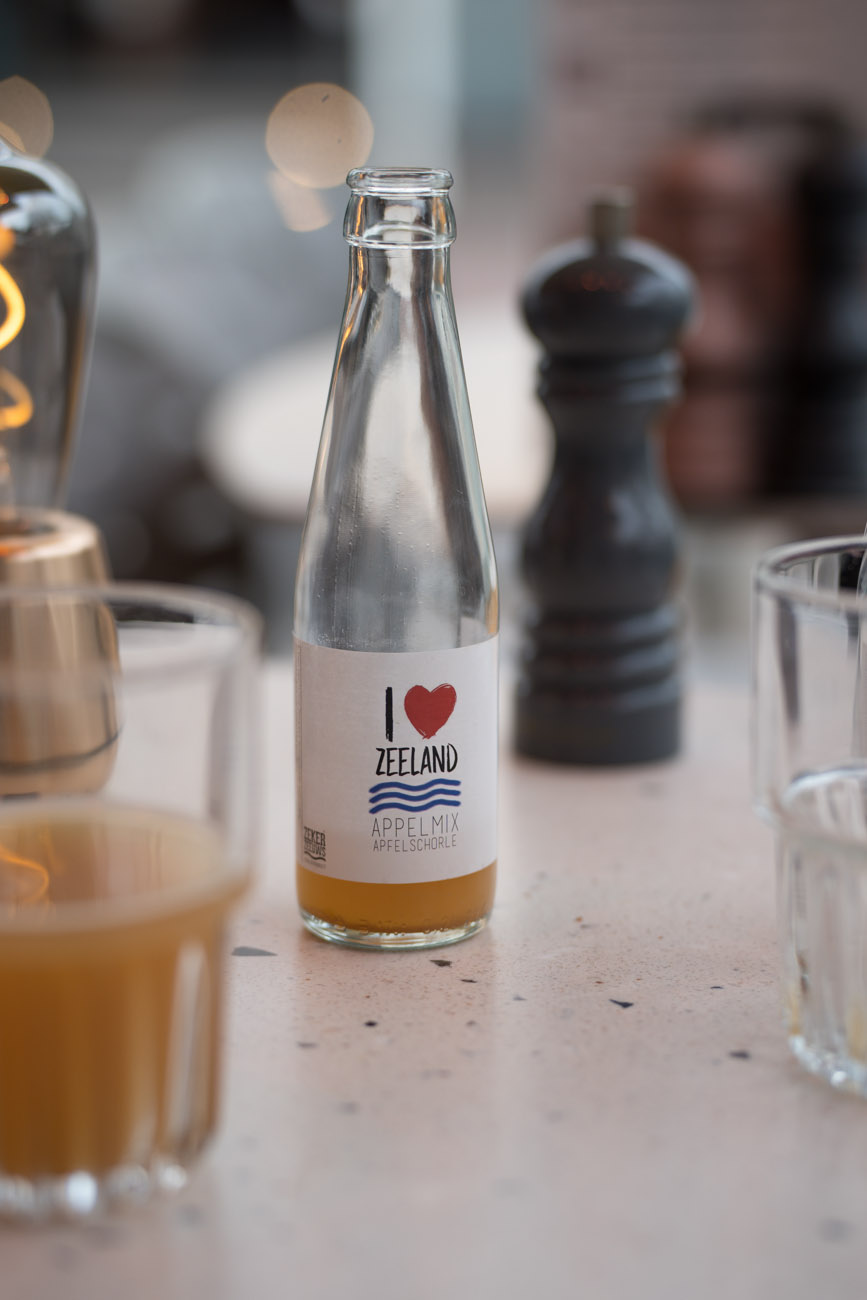 Ein Tisch mit einer kleinen Flasche Apfelschorle in der Mitte. Man sieht noch eine Pfeffermühle im Hintergrund und ein gefülltes Glas vorne.
