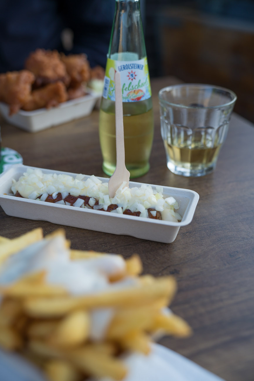 Ein Tisch mit Fastfood. In der Mitte sieht man ein Schälchen mit Friakndel Speciaal. Im Hintergrund steht eine kleine Flasche mit einem Glas Apfelschorle. 