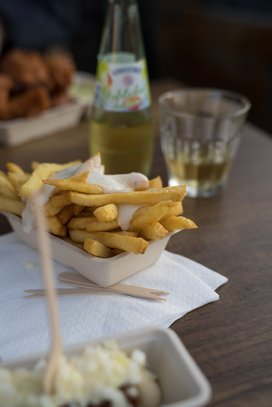 Ein Tisch mit Fastfood. In der Mitte sieht man ein Schälchen mit Pommes und einer weißen Soße. Im Hintergrund steht eine kleine Flasche mit einem Glas Apfelschorle. 