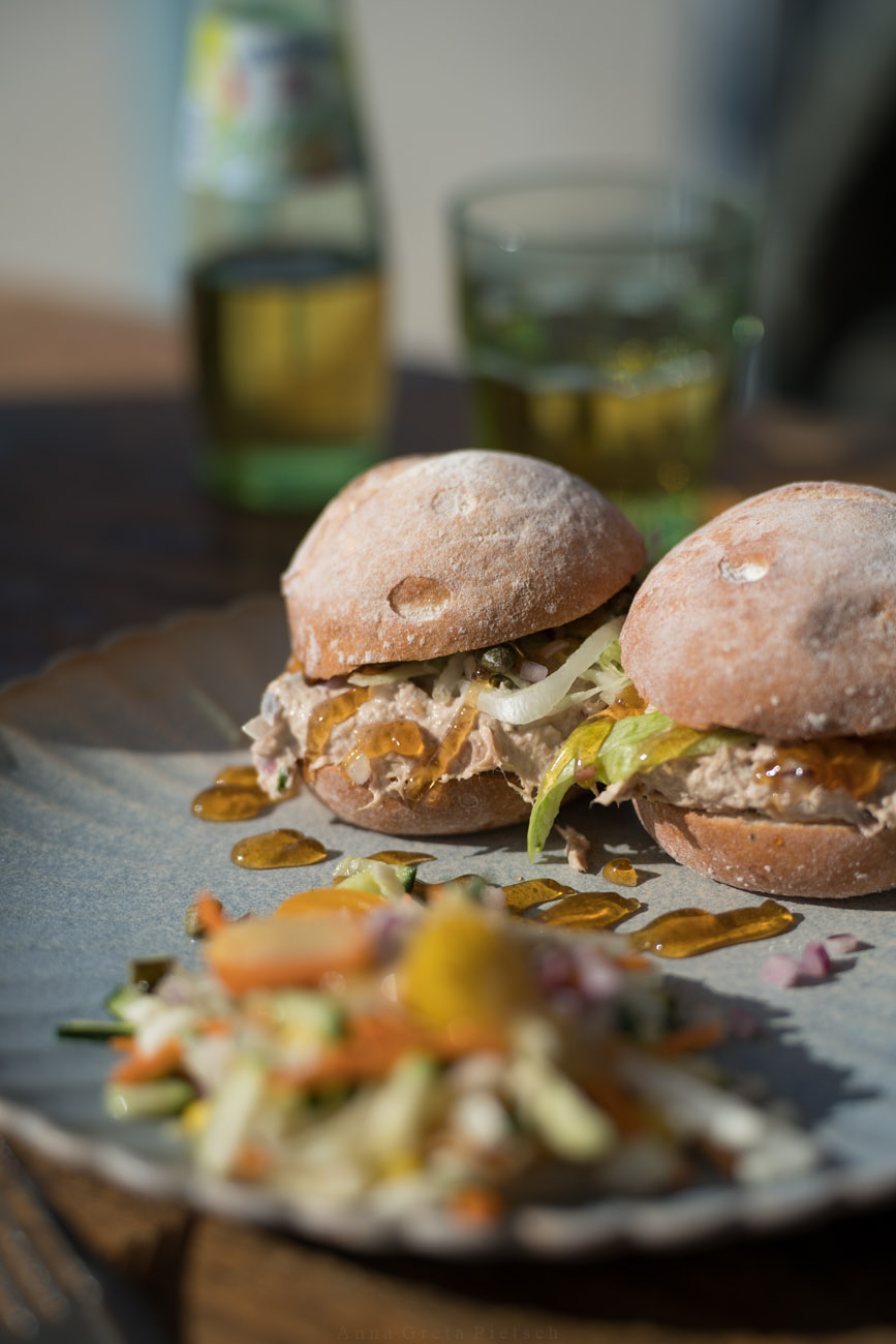 Zwei glutenfreie runde Brötchen auf einem Teller. Die sind mit einer Thunfischcreme und etwas Salat belegt. Man sieht im vordergrund unscharf den Anschnitt von einem Beilagensalat und Honig auf dem Teller.