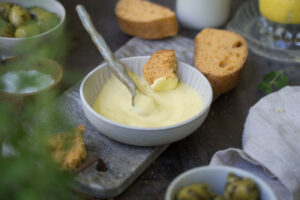 Selbstgemachte Aioli in einem Schälchen, Brotscheiben liegen drumherum und eine Flasche mit Milch ist im Hintergrund zu sehen. Im Vordergrund ist ein Schälchen mit Oliven.