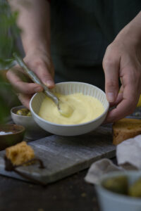 Selbstgemacht Aioli ohne Ei in eienr kleinen Schale. Zwei Hände halten diese. Ein Löffel aus Keramil (selbstgetöpfert) ist in der Aioli. Brotstücke liegen auf dem Tisch zum Dippen.