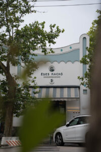 Edes Macko in der Außenansicht, ein weißes Gebäude mit hellblauen Verzierungen, Durch Blattgrün fotografiert, so dass einige Stellen unscharf sind