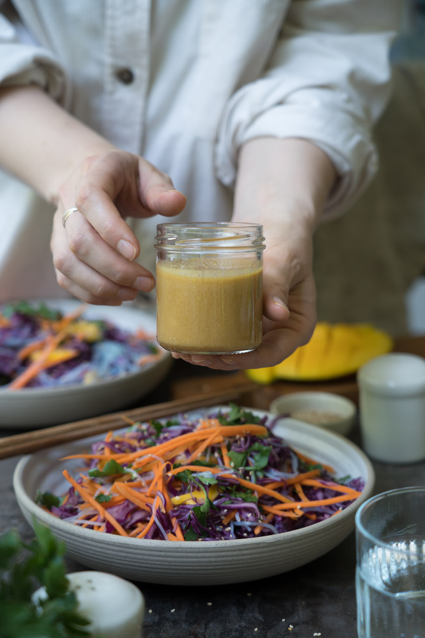 Erdnussdressing in einem Schraubglas, von zwei Händen gehalten. Auf dem Tisch sieht man Rotkohlsalat mit Glasnudeln und buntem Gemüse.