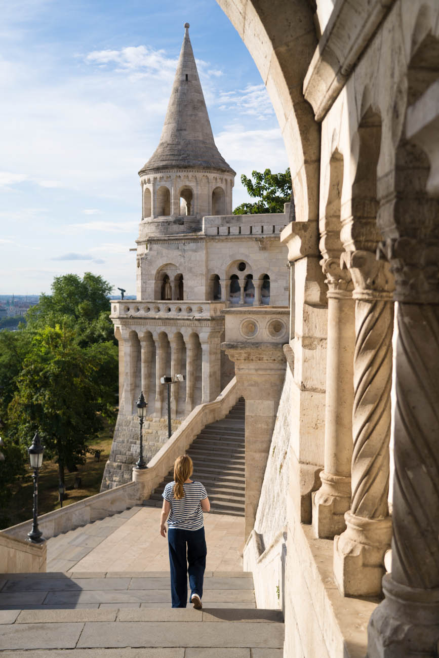 Die Fischerbastei am frühen Morgen in Budapest. Eine Person läuft eine Treppe herunter, im Hintergrund sieht man an Türmchen. Der Himmel ist blau mit einigen Wolken, die Sonne scheint.