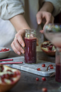 Selbstgemachter Granatapfelsirup in einer kleinen Glasflasche, steht auf einem kleinen Tablett aus Marmor und wird von einer Hand festgehalten/gerade abgestellt
