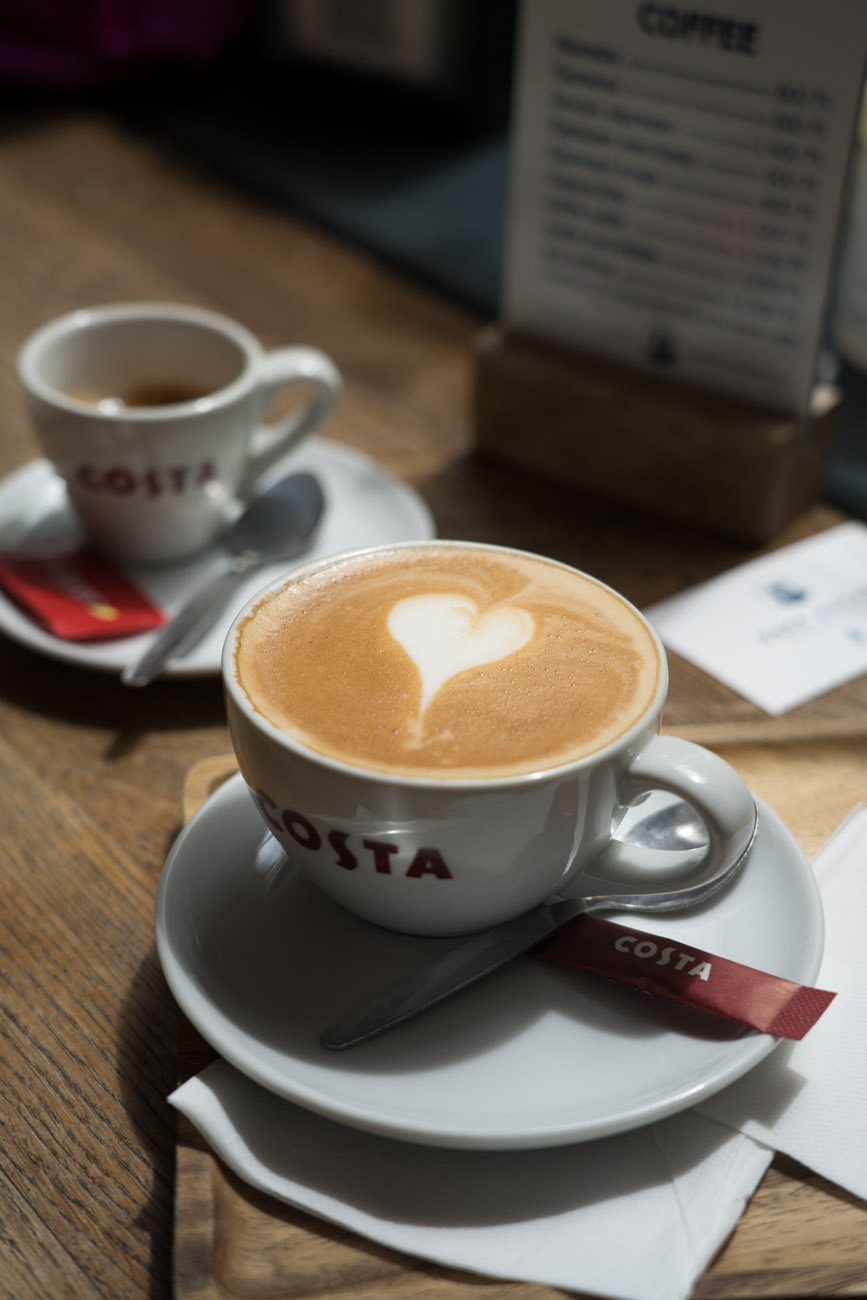 Eine große Tasse mit Cappucchino und einem Herz im Milchschaum, im Hintergrund steht eine kleien Tasse mit Espresso, auf einer Tischplatte aus Holz