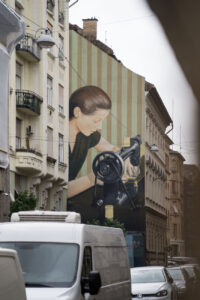 Eine Hauswand mit einem großen Kunstwerk/Murial, eine Frau mit einer Nähmaschine ist zu sehen