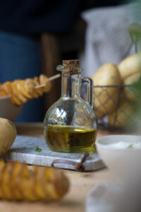 Olivenöl in einem kleinen Ölfläschchen mit Henkel und Kork-Stopfen auf einem Marmor-Tablett. Im Hintergrund sieht man Kartoffeln in einem Körbchen.