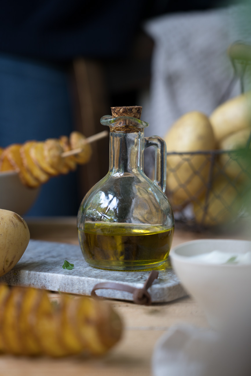 Olivenöl in einem kleinen Ölfläschchen mit Henkel und Kork-Stopfen auf einem Marmor-Tablett. Im Hintergrund sieht man Kartoffeln in einem Körbchen.