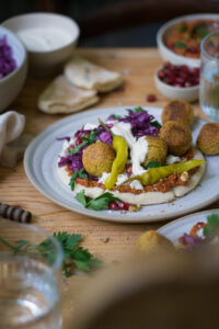 Pita mit Muhammara, Rotkohlsalat, Falafel, Peperoni und einem Dip auf einem beigen Teller. Es steht auf einem Holztisch.