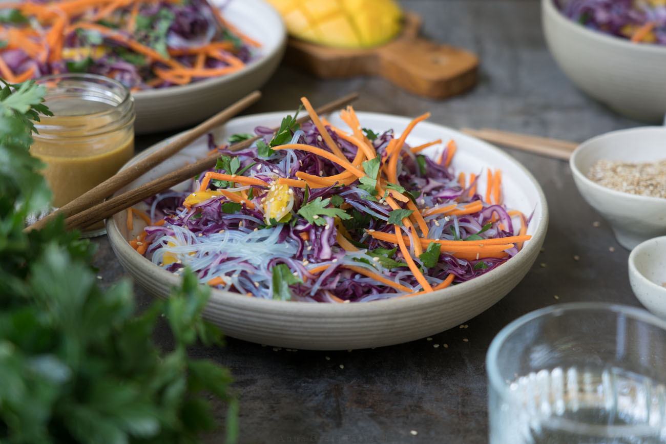 Rotkohlsalat mit Glasnudeln, fein geschnittenen Möhren, Petersilie und Mango in einer beigen Schale auf einem dunklen Tisch/Untergrund. Im hintergrund steht eine weitere Schale mit dem Salat. Essstäbchen aus Holz luegen bereit, ein Wasserglas und Salatdressing stehen auf dem Tisch.