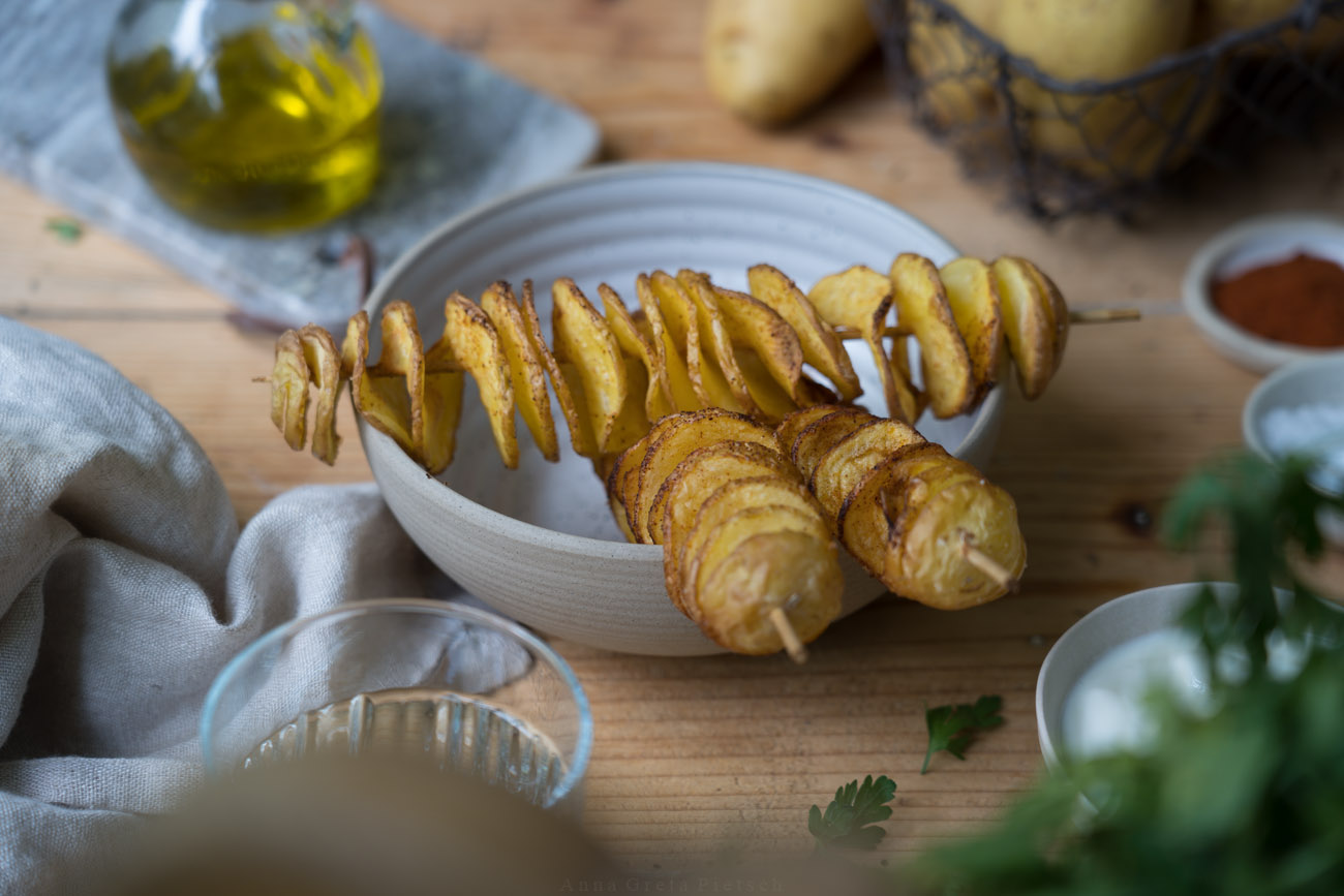 Eine Keramikschale mit drei selbstgemachten Spiralkartoffeln auf Holzspießen, im Hintergrund sieht man ein Ölfläschchen, Kartoffeln und im Vordergrund ein Glas Wasser.