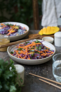 Rotkohlsalat mit Glasnudeln, fein geschnittenen Möhren, Petersilie und Mango in einer beigen Schale auf einem dunklen Tisch/Untergrund. Im hintergrund steht eine weitere Schale mit dem Salat. Essstäbchen aus Holz luegen bereit, ein Wasserglas und Salatdressing stehen auf dem Tisch.