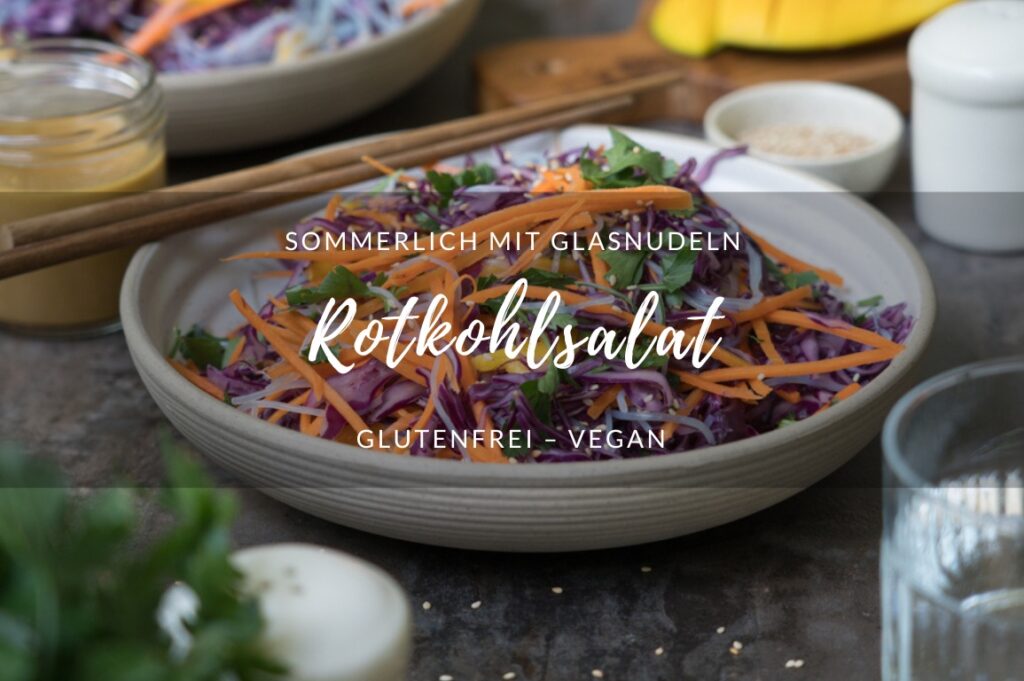 Rotkohlsalat mit Glasnudeln, fein geschnittenen Möhren, Petersilie und Mango in einer beigen Schale auf einem dunklen Tisch/Untergrund. Im hintergrund steht eine weitere Schale mit dem Salat. Essstäbchen aus Holz luegen bereit, ein Wasserglas und Salatdressing stehen auf dem Tisch.