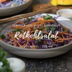 Rotkohlsalat mit Glasnudeln, fein geschnittenen Möhren, Petersilie und Mango in einer beigen Schale auf einem dunklen Tisch/Untergrund. Im hintergrund steht eine weitere Schale mit dem Salat. Essstäbchen aus Holz luegen bereit, ein Wasserglas und Salatdressing stehen auf dem Tisch.