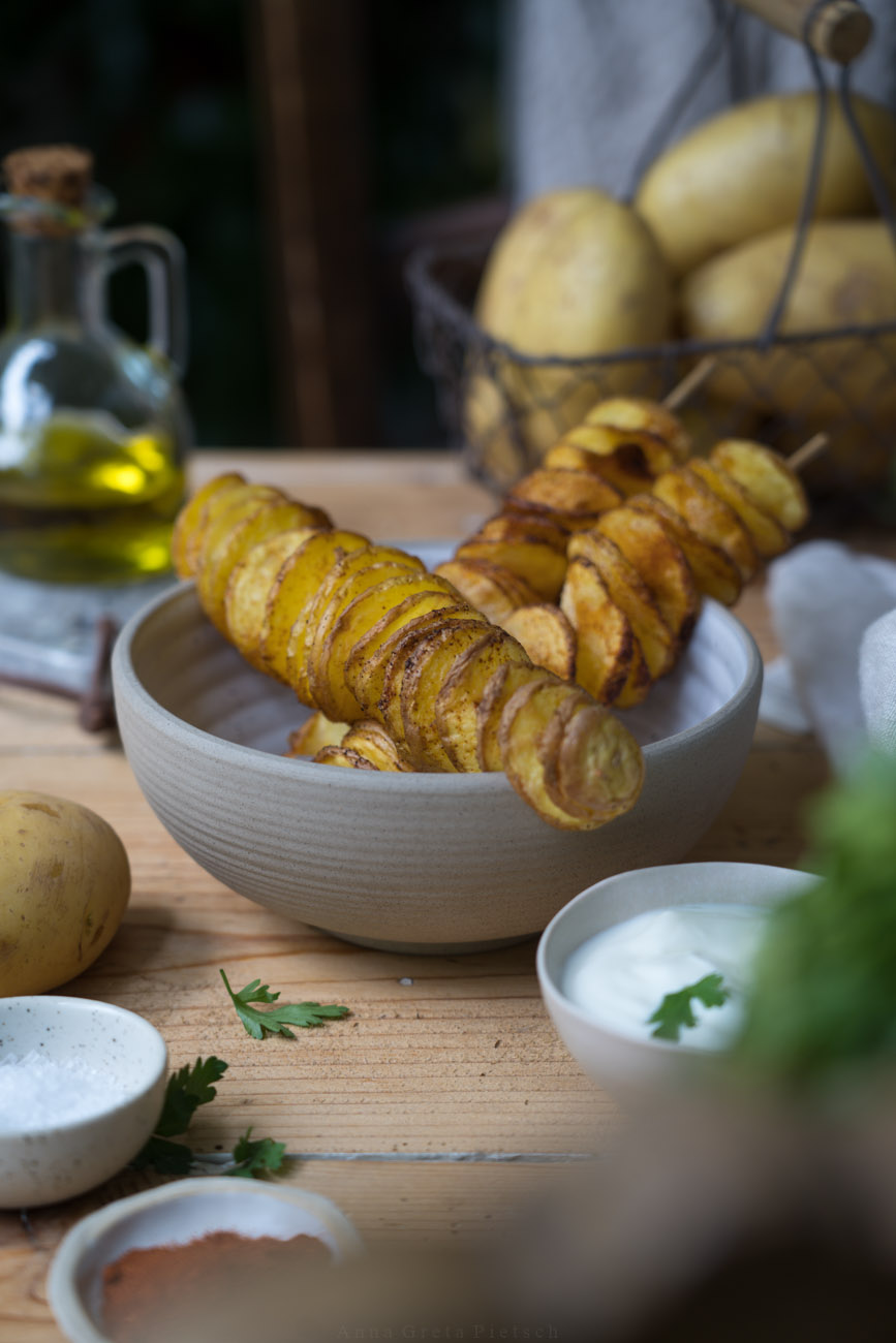 Spiralkartoffeln auf einem Schaschlikspieß in einer Keramikschale. Im Hintergrund sieht man ein Körbchen aus Draht mit Kartoffeln und Olivenöl in einer kleinen Flasche.