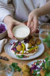 Ein Tahini-Joghurt-Dip in einem kleinen Keramik-Schälchen, gehalten von zwei Händen über einem gedeckten Tisch