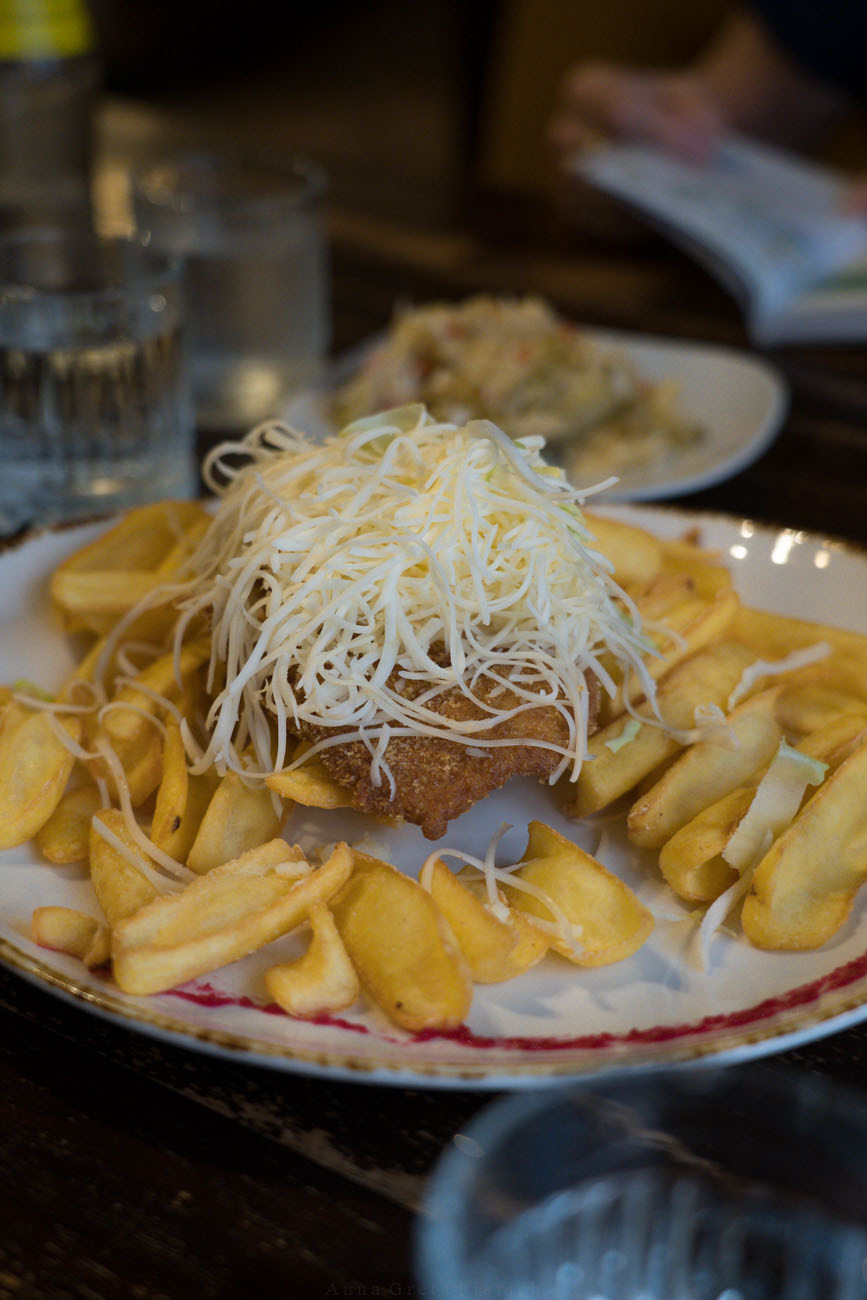 ein glutenfreies Cordonbleu auf einem Teller mit Pommes und viel geriebenem Käse obendrauf, steht auf einem Tisch, im Hintergrund ist eingelegtes Gemüse zu sehen