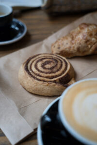 Eine Papiertüte auf einem Holztisch, darauf liegt eine flasche Zimtschnecke und ein Käsecroissant (glutenfrei). Im Vordergrund sieht man einen Cappuchino.