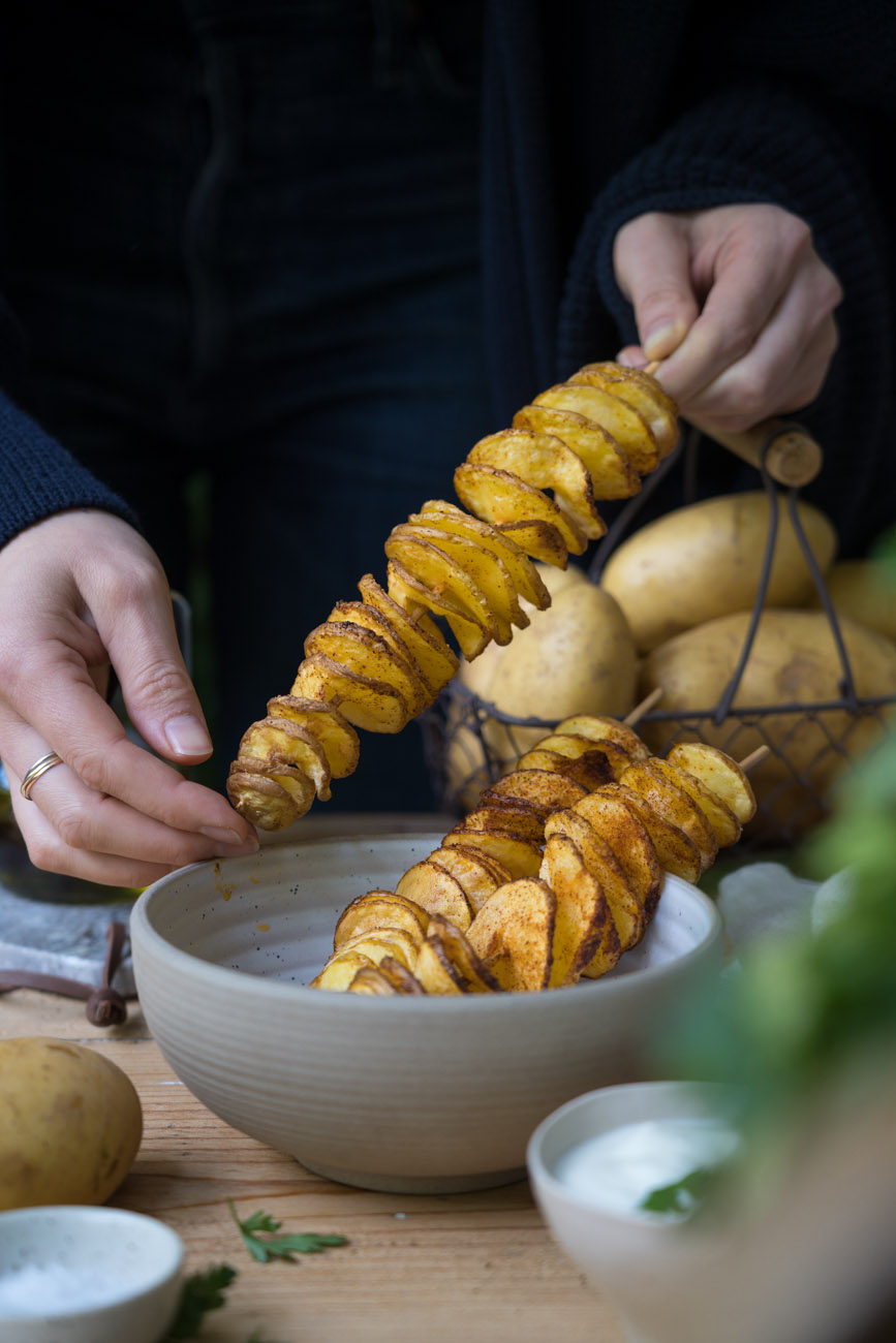 Zwei Hände halten einen Holzspieß mit Twister-Kartoffeln (spiralförmig geschnittene Kartoffeln), die in der Heißluftfritteuse knusprig gebacken wurden. 