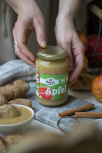 Ein Glas mit Apfelmus steht auf einem Tisch und wird von zwei Händen gehalten. Im Hintergrund sieht man Äpfel und Zimtstangen, Stockbrote.