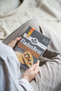 Zwei Hände halten eine Backmischung für glutenfreies Fladenbrot von Bauck über dem Schoß. Die Person trägt beige Kleidung.