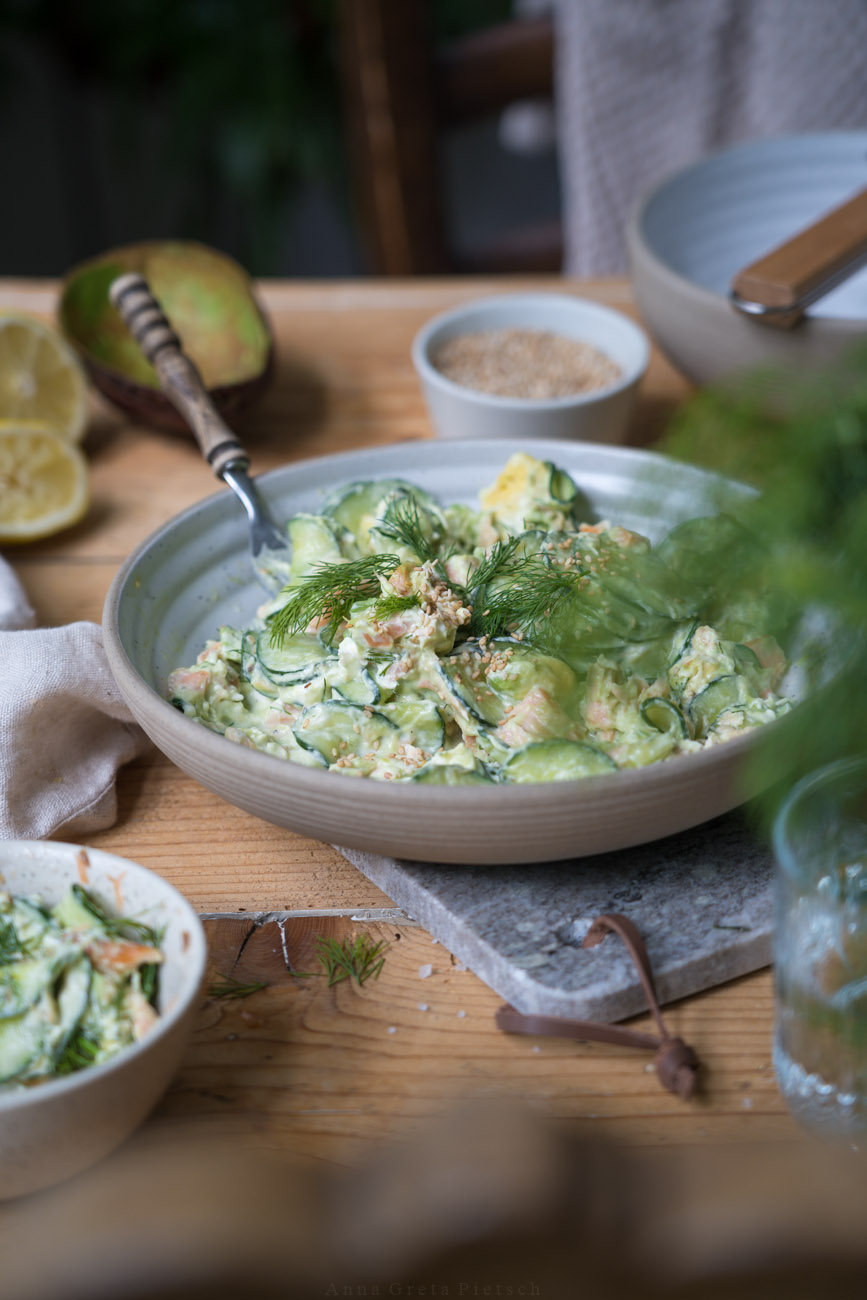 Ein Holztisch mit einem Marmorbrettchen, darauf steht eine Schale mit einem Gurkensalat mit Lachs.