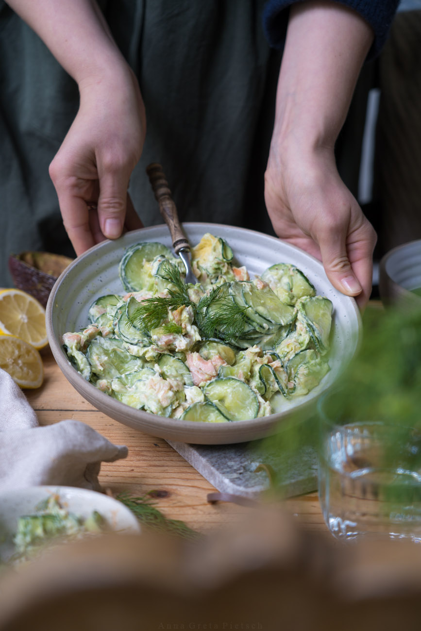 Gurkensalat mit KLachs und einer cremigen Soße aus Frischkäse in einer Schale, wird von zwei Händen auf einem Holztisch abgestellt.