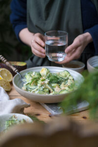 Ein Holztisch mit einem Marmorbrettchen, darauf steht eine Schale mit einem Gurkensalat mit Lachs, eine Person hält ein Wasserglas im Hintergrund.
