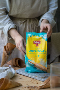 Eine Packung flutenfreies Mehl in Gelb und Türkis auf einem Marmorbretten, auf einem Holztisch. Im Hintergrund sieht man Eishörnchen.