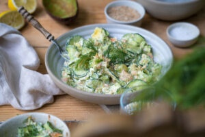 Ein Holztisch mit einem Marmorbrettchen, darauf steht eine Schale mit einem Gurkensalat mit Lachs, eine Person hält ein Wasserglas im Hintergrund.