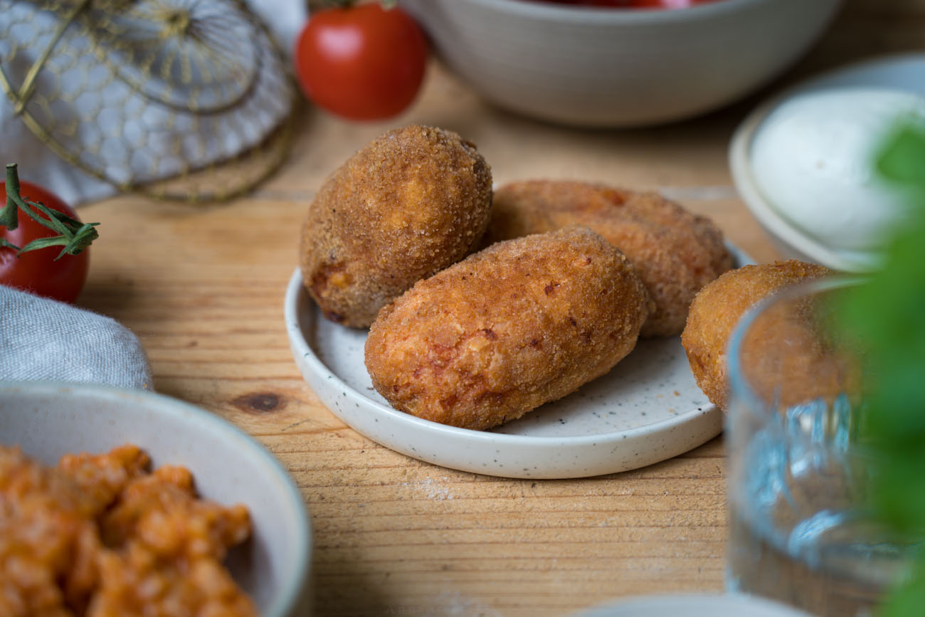 Suppli, italienische Reiskroketten. Drei Kroketten liegen auf einem kleinen Teller, eine auf dem Tisch dahinter. Die Tischplatte ist aus Holz. Man sieht unscharf Basilikum und eine Schale mit Risotto im Vordergrund.