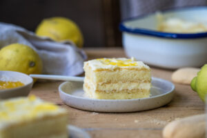 Ein Stück Zitronen-Tiramisu auf einem kleinen Teller, auf einem Holztisch Im Hintergrund sieht man eine Schale mit Tiramisu.