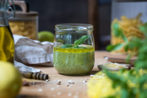 Ein Schraubglas mit Zitronen-Pesto, im Hintergrund steht ein Vorratsglas mit Pasta, eine Hand mit einem kleinen Löffel, der etwas Pesto entnehmen möchte, vorne erkennt man eine Zitrone und eine Ölflasche am Rand.
