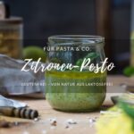 Ein Schraubglas mit Zitronen-Pesto, im Hintergrund steht ein Vorratsglas mit Pasta, eine Hand mit einem kleinen Löffel, der etwas Pesto entnehmen möchte, vorne erkennt man eine Zitrone und eine Ölflasche am Rand.