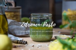Ein Schraubglas mit Zitronen-Pesto, im Hintergrund steht ein Vorratsglas mit Pasta, eine Hand mit einem kleinen Löffel, der etwas Pesto entnehmen möchte, vorne erkennt man eine Zitrone und eine Ölflasche am Rand.