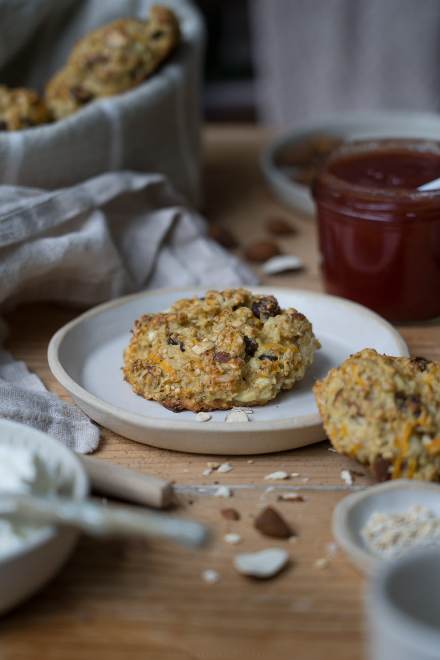 Ein Holztisch mit handgemachter, heller Keramit, eine Frühstücksszene, ein Brotkorb aus beigem Stoff, in der Mitte liegt ein Brötchen aus Haferflocken und Rosinen auf einem der Teller.