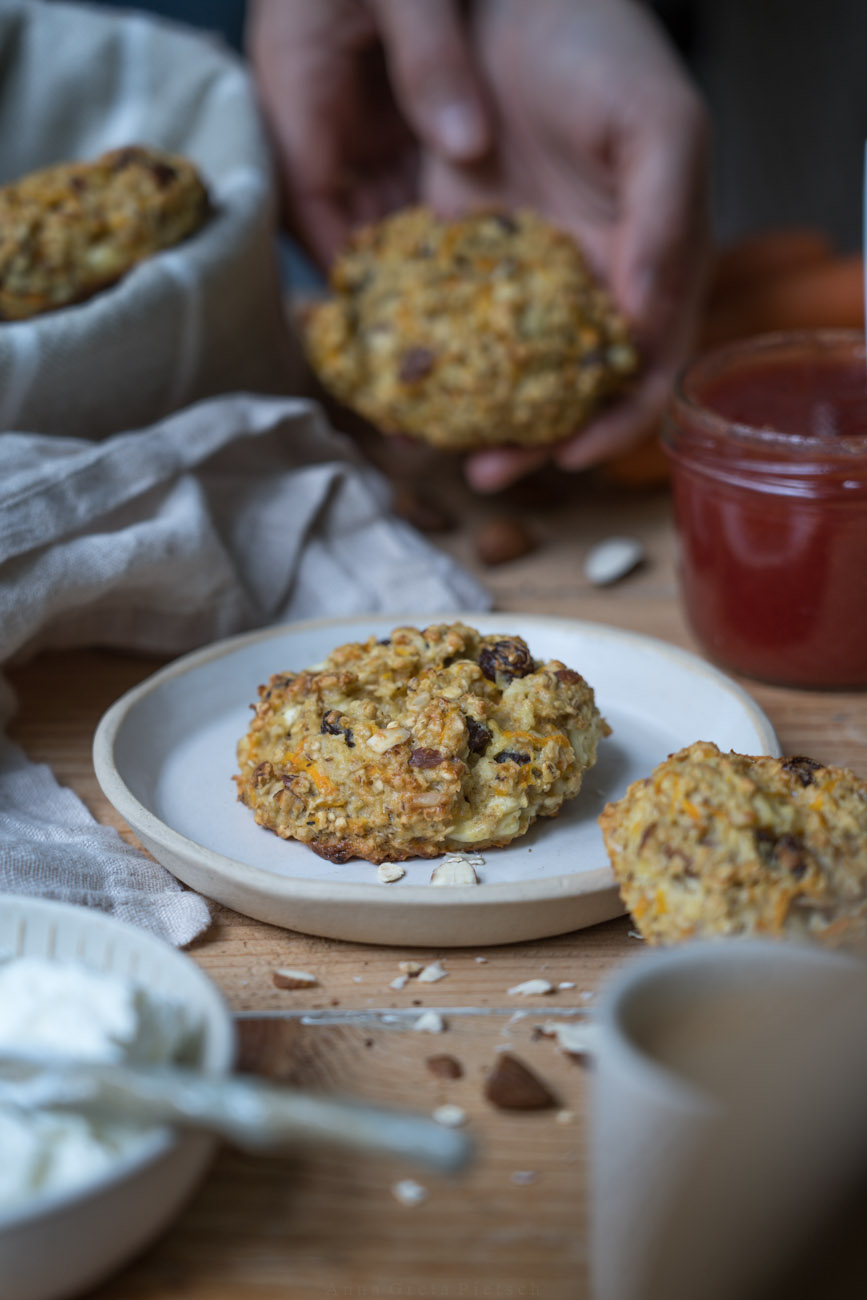 Ein Holztisch mit handgemachter, heller Keramit, eine Frühstücksszene, ein Brotkorb aus beigem Stoff, in der Mitte liegt ein Brötchen aus Haferflocken und Rosinen auf einem der Teller.