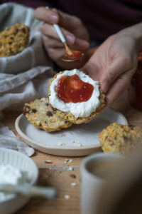 Ein Holztisch mit handgemachter, heller Keramit, eine Frühstücksszene, ein Brotkorb aus beigem Stoff, in der Mitte hält eine Hand eine Hälfte eines Brötchens aus Haferflocken und Rosinen und schmiert etwas Erdbeermarmelade darauf.