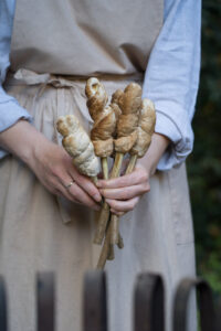 Eine PErson mit beiger Kleidung steht hinter einem Feuerkorb und hält mehrere Stäbe mit Stockbrot in den Händen.