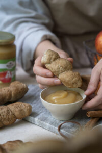 Eine Person hält ein Stockbrot in einer HAnd und dippt es in ein Schälchen mit Apfelmus. Die Person sitzt an einem Tisch. Man erkennt weitere Stockbrote.