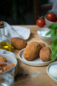Suppli, italienische Reiskroketten. Drei Kroketten liegen auf einem kleinen Teller, eine auf dem Tisch dahinter. Die Tischplatte ist aus Holz. Man sieht unscharf Basilikum und eine Schale mit Risotto im Vordergrund.