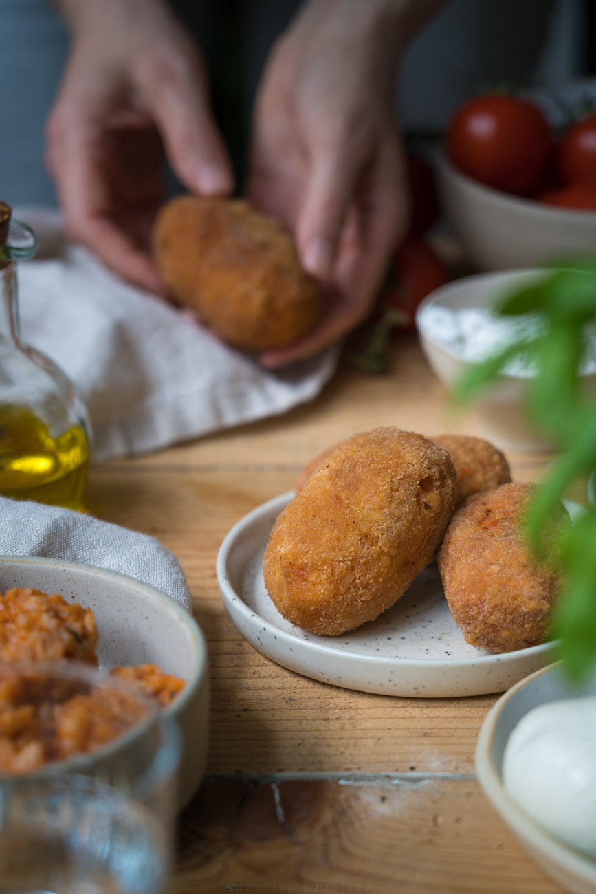 Suppli, italienische Reiskroketten. Drei Kroketten liegen auf einem kleinen Teller, eine wird dahinter von zwei Händen gehalten. Die Tischplatte ist aus Holz. Man sieht unscharf Basilikum und eine Schale mit Risotto im Vordergrund.